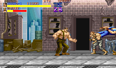 A shirtless fighter in tan pants stands in a defensive stance center-frame on a brick-walled street. Two armored enemies flank him—one to the left in a yellow outfit, one to the right in blue armor on a ledge. A wooden crate sits left of center. The HUD displays player health bars, score of 11890, and round timer at top left. Sprite-based fighters show crisp pixel art against a parallax brick background with shuttered windows and doorways.