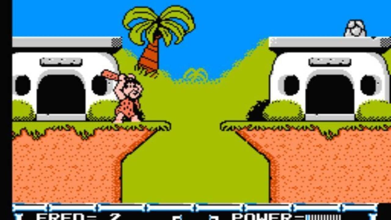 A pixelated side-scrolling level displays Fred Flintstone in a red and black spotted outfit standing on green grass near a white stone cave entrance on the left. A palm tree and blue sky occupy the upper center. On the right side, an identical cave structure sits across a green valley. Orange-brown rocky terrain fills the lower half, with a black status bar at the bottom showing health bars and power indicators in blue and white. The sprite uses 8-bit NES-era graphics with limited color palette.