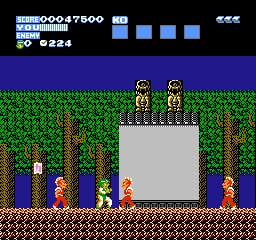 A side-scrolling gameplay scene features two sprite characters standing on a gray stone structure in the center. Below them are multiple colored enemies in red, blue, and brown positioned on brown brick ground. The background shows a green grass hill against a dark blue sky. A score display at the top left reads numbers in white text, with four blue health/status squares visible in the upper right. The scene uses a 16-bit NES color palette with distinct pixelated sprites and platform elements.