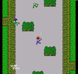A soldier sprite in blue uniform stands in the center of a gray road bordered by green grass and trees on both sides. Four dark green bush clusters are scattered across the vertical scrolling level at varying heights. The left edge displays "LIFE×0" in white text. The environment uses a top-down perspective with pixelated 8-bit graphics typical of NES-era arcade ports.