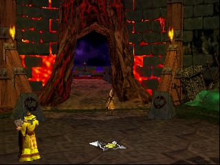 A yellow-clad character stands in a stone dungeon arena facing a large red demonic skull mouth structure with glowing orange and purple interior. The creature's form dominates the center background, with wooden torches flanking its sides. A defeated enemy lies on the ground near the player character. The environment uses warm earthen tones with stone textures, and the scene is lit by the skull's supernatural glow. Green vegetation is visible in the lower right corner of the arena.
