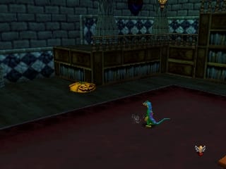 A gecko character stands on a dark red floor in the center of a tiled room with blue and green checkered walls and wooden bookcases. A glowing golden object rests on the floor to the left, and a small red enemy creature appears in the lower right corner. The scene uses a low-polygon 3D art style typical of Nintendo 64 games, with a fixed camera angle showing the interior space from an elevated perspective.