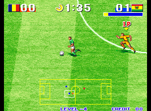 A soccer arcade game displays two pixelated player characters on a bright green grass field with a yellow goal frame visible at the bottom. The player in red controls the ball on the left side while an opposing player in yellow approaches from the right. The upper-left corner shows country flag icons and a score of 00, with a timer displaying 1:35. The upper-right displays a health or status bar alongside credit information marked 00. The bottom of the screen shows level designation and game status text. The sprite-based graphics feature low resolution typical of 1990s arcade games.