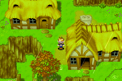 A top-down village scene rendered in 16-bit pixel art displays multiple thatched-roof cottages scattered across a bright green grass field. A small character sprite stands in the center of the landscape. Various building structures with brown roofing and yellow-toned wooden walls occupy the left, right, and lower portions of the screen. Trees and foliage appear throughout the scene in shades of green and brown. The color palette emphasizes vibrant greens and earth tones typical of Game Boy Advance graphics.
