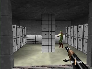 A first-person view shows a player character holding a rifle in the foreground, aiming into a gray concrete room. A central column made of stacked cubic blocks rises vertically in the middle distance. Filing cabinets line both the left and right walls in neat rows. An enemy soldier in yellow and brown clothing stands near the back of the room. The environment uses flat gray textures with basic geometric shapes, typical of N64-era 3D rendering with visible polygon-based architecture.