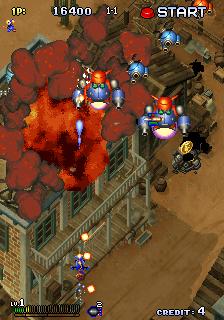 An isometric top-down arcade shooter displays a military compound with tan buildings and red-roofed structures. Multiple blue player aircraft with white markings are positioned across the center and lower portions of the screen, firing white bullet patterns upward toward explosions and enemy units. Orange fire bursts and smoke clouds fill the upper-center area. The UI shows a score of 16300 in the top-left corner with a START indicator, and a credit counter at the bottom. Tan and brown terrain with geometric building layouts creates the playfield's base layer.
