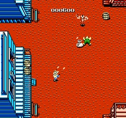 A bright red desert landscape displays a player character sprite in the lower-center area, with several enemy sprites scattered across the brown terrain. Blue and white building structures line the left edge of the screen. A score counter reading "000600" appears at the top center. The scene uses simple 8-bit pixel sprites against a solid red background, characteristic of NES-era graphics.