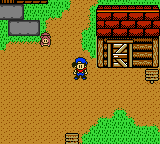 A top-down view of a farm area with a player character in blue clothing standing on tan-colored soil. A red wooden farmhouse with a chimney occupies the upper right. Green patches of grass and trees dot the landscape on the left and bottom edges. Brown fence sections are visible in the lower corners. The sprite-based graphics use a limited color palette typical of Game Boy Color hardware, with the scene displayed on a small screen resolution characteristic of the platform.