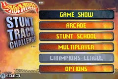 The title screen displays the Hot Wheels Stunt Track Challenge logo in yellow text at the top left, with a flaming orange and red gradient background. Below it are five menu options arranged vertically in tan and gray colored buttons: Game Show, Stunt School, Multiplayer, Champions League, and Options. A Select indicator appears at the bottom of the screen. The overall color scheme uses warm oranges, yellows, and earth tones typical of Game Boy Advance graphics.