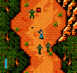 A top-down view of a sandy terrain surrounded by dense green vegetation. Multiple pixel-art soldiers in blue uniforms are scattered across the orange-brown ground, with one prominent figure in the center. Small green trees and brown rock formations dot the landscape. A blue health or ammo meter appears on the left edge of the screen. The sprite-based graphics use a limited NES color palette of greens, oranges, and earth tones typical of early 1990s action games.