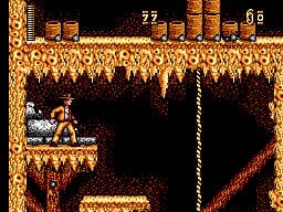 A character in brown clothing stands on a narrow platform on the left side of the screen against a dark background. Golden-brown textured walls fill the upper half of the image, suggesting a cave or temple interior. The score display shows 22 at the top left and 6 at the top right. A rope or vertical element hangs in the center-right of the screen. The sprite work uses orange and brown tones with black outlines typical of early SMS graphics.