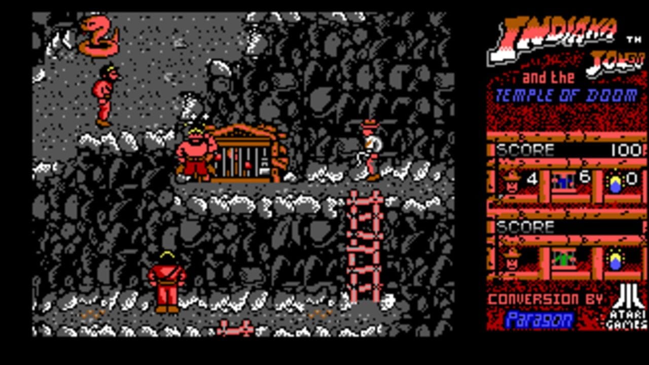 An Atari 8-bit platformer level displays Indiana Jones in red clothing on a gray stone temple interior with skull-patterned walls. Multiple enemies in red appear at various heights—one on a high ledge, one mid-platform, and one on the ground level. A small red building structure sits in the center-right area. The right panel shows the game title, score of 100, a lives counter showing 2, and inventory slots displaying collected items in different colors. The low-resolution pixelated graphics use a limited palette of red, gray, black, and white.