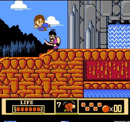 A player character stands on a brown brick platform in the center of the screen, with another character visible on the upper left near a white cloud and blue sky. A brick structure with red decorative elements occupies the right side. The bottom UI displays a health bar labeled "LIFE" on the left, a number "7" in the center, and an orange item counter showing "x00" on the right. The scene uses 8-bit NES sprite graphics with a limited color palette of reds, browns, blues, and flesh tones typical of early 1990s console games.