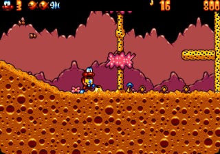 A side-scrolling platformer level displaying a small character sprite positioned on an orange-brown terrain with circular crater patterns. Pink spiky enemy creatures are visible in the middle ground, with golden platform structures extending upward on the right side. The background shows layered mauve-colored hills creating a parallax depth effect. The top of the screen displays a score of 880 and the number 16, with pixelated UI elements in the upper corners. The overall color palette uses warm earth tones for the ground and cool purples for the background, typical of 16-bit sprite-based graphics.