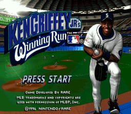 A baseball player in a white uniform crouches in a batting stance on a green baseball diamond, positioned in the center-right of the screen. Behind him, a blue scoreboard and stadium structure are visible. Large white text reading "WINNING RUN" appears above the player in a stylized font. A smaller "PRESS START" prompt is displayed in the lower portion of the image. The lower left corner contains small developer credits text. The overall color palette features bright greens, blues, and whites typical of a 16-bit sprite-based sports game.