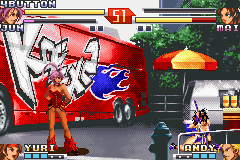 A fighting game battle unfolds on a street scene with a red fire truck visible in the background. Two characters face off in the center, with red-hued effects suggesting an active attack or special move. The top of the screen displays health bars and character portraits in the upper left and right corners. The bottom left shows player information and icons, while the bottom right displays opponent details. The sprite-based characters and pixelated background elements are rendered in typical Game Boy Advance resolution, with a daylit urban setting featuring green foliage and industrial structures behind the fighters.