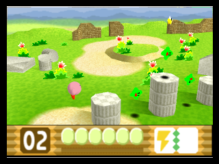 A 3D level viewed from an isometric perspective shows Kirby as a pink spherical character in the center of a grassy field dotted with brown barrels, white cylindrical structures, and red flowers. Mountains and stone buildings appear in the distant background under a clear sky. The HUD at the bottom displays level 02, six circular life counters, and a power indicator. The art style features pre-rendered 3D environments with vibrant green grass and simple geometric terrain elements.