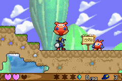 A side-scrolling platformer level displays Klonoa, a small red and white character with pointed ears, standing on a green platform in the center of the screen. A large green vertical structure rises behind him against a purple sky with white clouds. Brown rocky terrain forms the level ground, with blue water visible in the lower left. The HUD shows two red heart icons on the left, a brown bar divided into segments at the bottom left, and numeric values on the bottom right. The art style uses colorful 16-bit sprites with detailed pixel animation against a parallax background.
