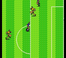 A soccer field viewed from above displays bright green pitch with white field markings including a center circle and goal lines. Two teams of pixelated players, distinguished by red and blue coloring, are positioned across the field. One player holds the ball near the center, while teammates and opponents spread across the pitch. The NES-era sprites are small and blocky against the solid-colored background with minimal detail.