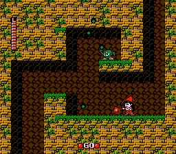 A top-down maze-like level displays a player character in the center standing on a brick-textured floor surrounded by dark stone walls forming corridors. A green enemy sprite appears in the upper-left passage, while a red object labeled "GO" sits below the player. The surrounding walls are constructed from brown and red brick tiles, creating an L-shaped pathway against a tan and gold brick exterior background. Yellow and green grass textures border the outer edges of the level layout.
