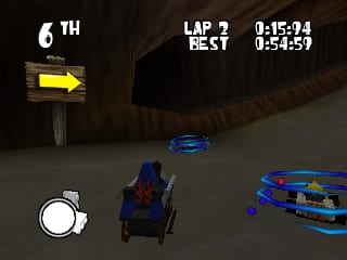 A blue LEGO race car is positioned in the center of a dark tunnel-like track, with another colorful vehicle visible to the right. The HUD displays "6TH" position in the upper left, lap information showing "LAP 7 BEST" with timer readings "0:15:04" and "0:54:59" in the upper right. A directional arrow sign points left on the track wall. The lower left corner contains a circular minimap or radar interface. The 3D polygon-based graphics show a indoor cavern setting with brown rocky walls and realistic lighting effects typical of N64-era racing games.