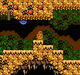 A multi-layered 2D platformer level displays a yellow stone structure with crenellated walls in the center-right, surrounded by brown earth and green foliage. A small blue character stands on a platform in the upper portion of the screen. Sharp brown and tan spikes protrude from the ground below the stone structure. The background shows dark black void areas with scattered golden coin or gem sprites filling much of the upper screen. The NES-era pixel art uses warm earth tones, gold highlights, and a limited color palette typical of 8-bit graphics.