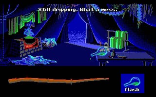 A dark cave scene rendered in EGA-palette blues and greens displays a figure standing on a central platform surrounded by glowing mushrooms, crystals, and geometric structures. A wooden staff is prominently displayed in the center-lower portion of the screen. The top of the screen shows text reading "Still dripping. What a mess." The Flask logo appears in the bottom-right corner, and the entire scene uses a low-resolution pixelated art style typical of early 1990s MS-DOS adventure games.