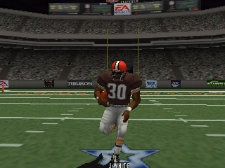 A player character wearing an orange helmet and brown jersey with number 30 stands at midfield in a football stadium, holding the ball. The field stretches behind with yard markers visible, while a full crowd fills the stands under stadium lights. A scoreboard is visible at the top center of the stadium. The 3D graphics show low polygon count and texture detail characteristic of early 2000s N64 sports games.