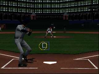 A batter in gray uniform stands at home plate in batting stance, viewed from behind. The pitcher is visible on the mound in the distance, with the catcher crouched behind home plate. A green targeting reticle appears in the lower-center area of the screen. The stadium features a dark green outfield wall with scoreboard displays in the upper background. The infield dirt and baseline are rendered in brown tones, with bright white foul lines visible. The graphics show the polygon-based 3D style characteristic of N64-era baseball games with moderate texture detail and flat-shaded surfaces.