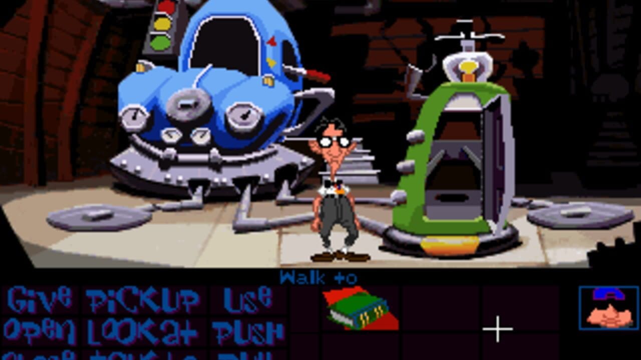 A pixelated scene shows a brown-haired character in a brown outfit standing in an industrial space with a blue UFO on the left featuring control dials and antennae, and a green machine on the right with a helicopter blade on top. A traffic light with red, yellow, and green lights is visible in the upper left. The floor is tan with circular metallic panels. At the bottom, blue text displays interface commands including "Give," "Pickup," "Open," "Look," and "Push," along with a small inventory icon in the lower right corner showing a character portrait.