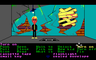 A low-resolution EGA graphics scene displays a character in dark clothing standing before a colorful teal interior with red brick structures and yellow horizontal elements overhead. The bottom of the screen shows a dark command menu with green text listing interactive verbs: "Turn on", "Open", "Talk to", "Unlock", and partial options for cassette tape and other inventory-related commands. The interface indicates a point-and-click adventure game structure with text-based interaction options for puzzle-solving.