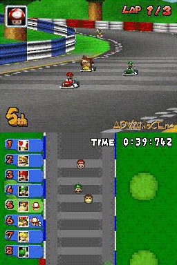 A Mario Kart DS race displays three karts on a gray asphalt track with white lane markings and green grass borders. The HUD shows lap 9/3 in the top-left, race time 0:39:762 in the center, and a vertical driver roster on the left side with character portraits. The track curves ahead with blue and red guardrails visible in the background. A bright green item box icon appears in the lower-right corner. The view is from behind the lead racer's perspective during active gameplay.