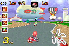 Mario Kart: Super Circuit gameplay showing a race in progress on a track with water. The HUD displays lap 3/3 at top left, with item and character icons in the upper left corner. The player's kart, piloted by what appears to be Toad, occupies the center-bottom of the screen in red. A second-place indicator and character portrait appear in the lower right. The track has grass borders and water obstacles, rendered in bright colors typical of GBA graphics. A finish line banner with Mario Kart branding is visible in the distance.