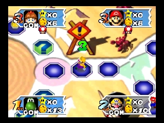 A Mario Party 3 minigame board displays four player portraits in the corners—Mario top-left, Peach top-right, Luigi bottom-left, and Daisy bottom-right—each labeled with "XO" indicators. The center shows a tan-colored game space with a red and yellow rotating wheel mechanism in the middle, surrounded by seven blue circular button icons arranged in a pattern. The 64-bit sprite graphics and bright primary color palette are typical of early-2000s Nintendo 64 presentation.