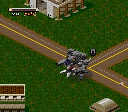 A mech unit with a boxy, industrial design stands at a crossroads on a bright green terrain, viewed from an isometric angle. The mech is rendered in dark grays and metallic tones with visible weapons mounted on its frame. A dirt road creates a cross pattern across the grassy field. The top-left corner displays a health/status bar and unit information. A small overhead map thumbnail appears in the bottom-left corner showing the surrounding area layout. The sprite-based graphics use a limited color palette typical of SNES-era games.