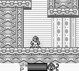 Mega Man stands in the center-left of a monochrome Game Boy screen, facing right in a detailed industrial interior with ornate pillars, horizontal paneling, and decorative rectangular patterns on the walls. The character sprite is small and pixel-based, rendered in black against a light background. A small circular object appears in the lower right corner, likely a weapon or item. The level design shows multi-layered architectural elements with repeating geometric motifs typical of 8-bit Game Boy graphics.