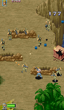 A top-down isometric view of a sandy desert level populated with multiple soldier sprites scattered across the terrain. The player's character appears in the upper-right area with a health bar displayed above. Rocky outcrops and palm trees are visible in the lower portion, with a pink building structure on the right edge. The HUD shows a score of 177 in the upper-right corner and health/ammo indicators at the bottom. The color palette consists of tan sand, brown rocks, and green vegetation against a 16-bit sprite-based aesthetic typical of early 1990s arcade action games.
