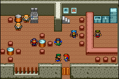 A top-down view of an indoor facility with tan-colored tile flooring shows multiple characters and objects scattered across the space. Several armored or uniformed figures stand in various positions, some near brown crate-like objects and a central reddish structure. A blue tank or vehicle sits in the upper-right area. Shelving units line the walls, containing boxes and equipment. The right side features a separate enclosed room with storage shelving. Wooden floorboards and concrete barriers form the room's boundaries. The pixel art uses a muted brown and blue color palette typical of early 2000s Game Boy Advance graphics.