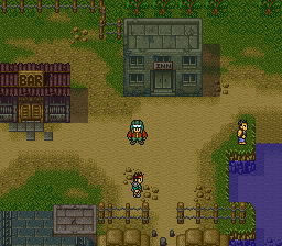 A top-down view of a sandy village area displays multiple characters and structures. A player-controlled character stands in the center, with two additional characters visible nearby. A wooden building with a gray roof occupies the upper left, while a larger gray stone structure with a door sits at the upper center. The scene includes patches of green grass, wooden fences, trees, and what appears to be water in purple tones on the right edge. The pixel art uses an isometric perspective typical of SNES-era RPGs, with a warm earth-tone color palette for the terrain.