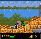 A small sprite-based soldier character stands on a tan-colored rocky terrain in the center-left of the screen, facing right. Behind him sits a yellow and orange tank. A blue sky with white clouds fills the upper portion, with green trees visible along the horizon line. The HUD displays green "LIFE" text with a health bar on the lower left and "H 00" in yellow on the lower right. The pixelated art style and color palette are typical of Game Boy Color graphics from 1999.