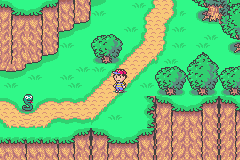 A top-down view of a vibrant green forest area with brown brick-textured ground paths winding horizontally across the screen. Two trees with green foliage dot the landscape on the right side, while brown-tiled cliff or building formations line the upper left and lower portions. A yellow-brown character sprite stands on the central path. The scene uses a 16-bit pixel art style with bright, saturated colors typical of early GBA graphics.