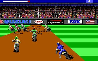 A motocross racing scene displays a dirt track with brown terrain and green grass sections. A blue-clad motorcycle rider on the right approaches a jump while several opposing riders in red and green occupy positions on the track. A crowded stadium with spectators fills the background. Sponsor logos including Suzuki, JCB, and EJK appear on a blue banner at the top. The color palette is bright with blocky pixelated graphics typical of late 1980s DOS games. The track layout suggests a top-down perspective view of the racing action.