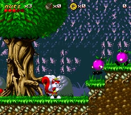 A red squirrel character stands on a brown platform in the lower-left area of a colorful side-scrolling level. The background features a large brown tree trunk on the left, green foliage at the top, and a blue starry night sky. Enemy creatures and projectiles scatter across the scene, with green mushroom-like obstacles on the right side and brown terrain at the bottom. The HUD displays "NUZ" with a health counter reading 3, a coin count showing 0, and additional score indicators in the top-left corner. The sprite-based graphics use a bright 16-bit color palette typical of SNES-era platformers.