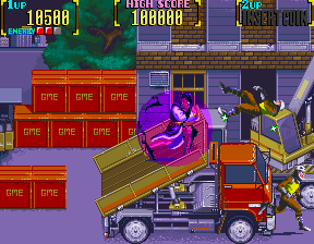 A side-scrolling beat-em-up scene displays two muscular fighters in combat stance on a wooden platform near an oversized red truck. The left fighter wears purple clothing while the right fighter is dressed in yellow and black. Behind them, a warehouse interior features stacked brown crates arranged in a grid pattern and purple-toned walls. The HUD shows score displays at the top left and right edges. The sprite-based graphics use a bright palette of purple, red, orange, and brown colors typical of early 1990s arcade hardware.