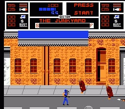 A blue-clad player character stands on a gray street in front of an orange brick building with multiple arched doorways and windows. A red object appears to the right of the character. The top of the screen displays a multi-row HUD with score information, level indicators reading "PRESS START", and "NARC" branding. A blue horizontal bar and checkered pattern border separate the play area from the interface. The sprite-based graphics use a limited color palette typical of NES-era arcade conversions.