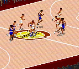 A basketball court viewed from an isometric angle shows ten players in two teams—one wearing blue jerseys, the other in tan—positioned around the center court logo. The wooden court features painted white lines marking boundaries and the center circle. Players are rendered as small sprites with visible pixelation typical of SNES-era graphics. The court's perspective creates depth with the far end appearing smaller than the near end. The color palette consists of warm wood tones, bright blues, tans, and the team logos visible on the court floor.