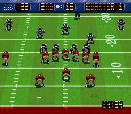 A football field viewed from above displays two teams lined up in formation. The upper team wears dark uniforms with white numbers, positioned in a defensive setup across the field. The lower team wears red uniforms, arranged in an offensive formation near the line of scrimmage. White yard line markers and numbers are visible on the green field. The top of the screen shows a dark blue scoreboard with game information and team icons. The bottom right corner displays a small stat panel with white text and numbers. Pixelated 16-bit sprite graphics represent all players and field elements.
