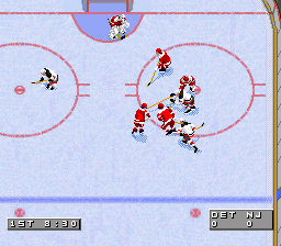 A hockey game in progress on an ice rink with a light blue background. Two teams of players in red and black jerseys are clustered around the center ice area near the goal crease. Pink circle outlines mark defensive zones on the ice. The scoreboard displays in the upper corners show game statistics. Sprite-based players are rendered in low resolution typical of SNES graphics, with simple pixelated animations. The rink has blue boards along the edges and the overhead view shows the full width of the playing surface.