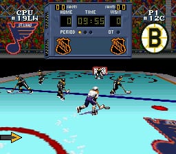 A hockey match viewed from above shows a light blue ice rink with two teams of pixelated players positioned across the surface. The scoreboard at the top displays the game time, period, and score between two teams. Team logos appear in the upper corners, with the Boston Bruins logo visible on the right. Red and blue player sprites are scattered across the rink, with a puck visible in play. The rink boundaries are outlined in dark colors, and the sprite-based graphics typical of early 1990s SNES sports games are evident throughout the scene.