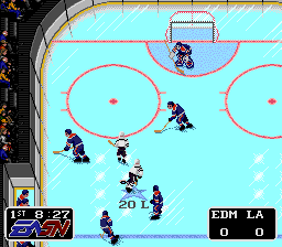 An overhead view of a hockey rink with a light blue playing surface divided by red lines and circles. Two teams of pixelated players in dark and light jerseys are positioned across the ice, with one player holding the puck near center ice. A goal net is visible at the top of the screen. The UI displays "EDM LA" in the lower right corner, indicating Edmonton versus Los Angeles. Score and game information appear in small text panels on the left side of the screen. The sprite-based graphics and color palette are characteristic of early 1990s SNES sports games.